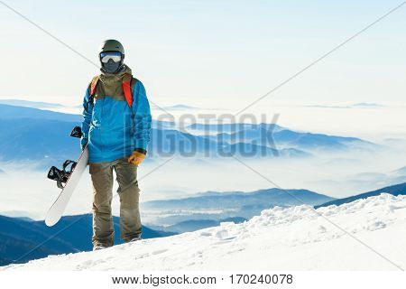Young Snowboarder In Helmet Standing At The Top Of A Mountain With Beautiful Sky On Background