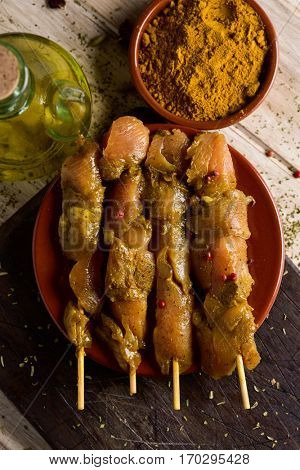 closeup of some raw spiced chicken meat skewers in an earthenware plate, on a rustic wooden table, next to a glass cruet with olive oil and a bowl with curry powder