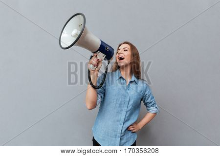 Woman in shirt screaming in megaphone with arm at hip in studio. Isolated gray background