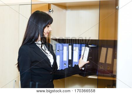 Beautiful young brunette woman take a folder with documents from cabinet in office.