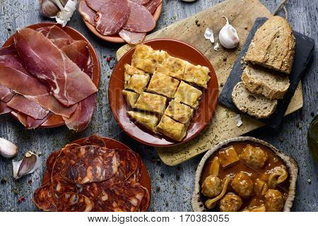 high-angle shot of some plates with an assortment of different spanish cold meats, such as serrano ham, chorizo or cured pork tenderloin, a spanish omelette and meatballs with cuttlefish, on a table