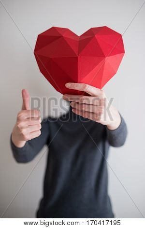 Woman holding red polygonal paper heart shape covering her face and gesturing thumb up. Shallow depth of field