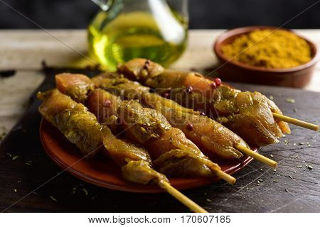 closeup of some raw spiced chicken meat skewers in an earthenware plate, on a rustic wooden table, next to a glass cruet with olive oil and a bowl with curry powder