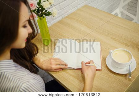 Pretty smiling woman makes entry in her diary on table. One hand writes notes and other holds skraping book. Cup of cappuccino, vase with pink and white roses stand on desk.