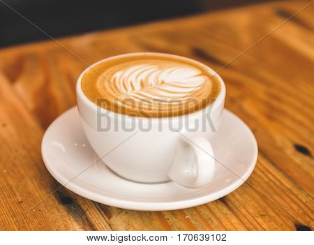 A carefully prepared cappuccino in a simple white cup and plate sitting on wooden table.