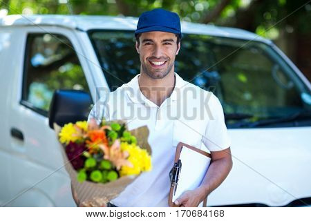 Portrait of smiling delivery man holding flower bouquet and clipboard while standing by van