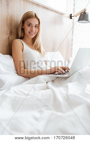 Vertical image of woman on bed with laptop and looking at camera. Side view