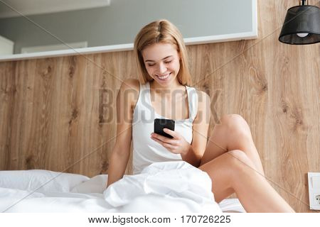Smiling woman using phone and sitting on bed in bedroom