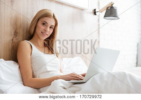 Side view of pretty woman lying on bed using laptop and looking at camera