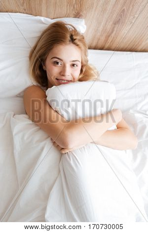 Top view of woman lying on bed, embracing with pillow and looking at camera. Vertical image