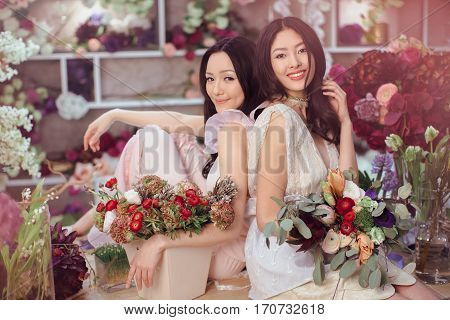 Fashion models in tender dresses posing and looking at camera. Beautiful asian florist girls making bouquet of flowers on table for sale against floral bokeh background in flower shop indoors. Two attractive asian females florists working in store.