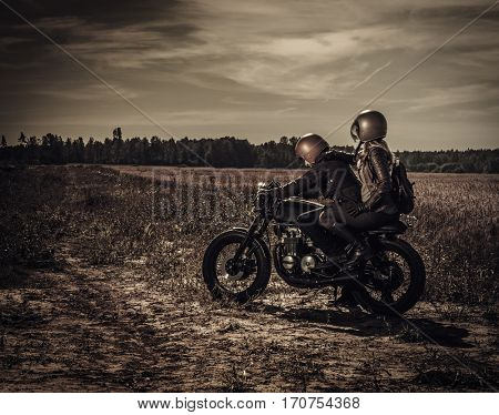 Young, stylish cafe racer couple on vintage custom motorcycles in field.