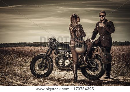 Young, stylish cafe racer couple on vintage custom motorcycles in field.