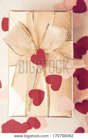 Overhead shot of a gold gift box tied with ribbon to a bow and scattered with red and pink confetti love hearts.
