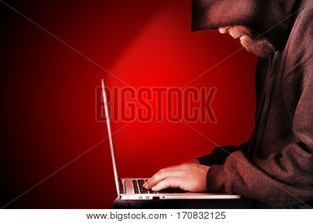Male computer hacker wearing a hooded top leaning over a laptop with red warning background. The screen light illuminates the man with a beard performing illegal activities. Graduated copy space on the left