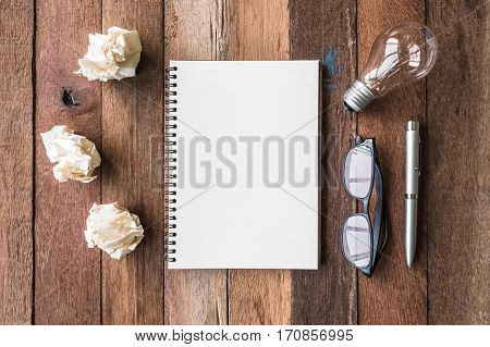 Top view of notebook with crumpled paper balls pen glasses and light bulb on wooden table background.