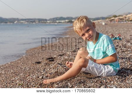 Happy smiling small boy sitting on the beach and playing with stones. Hands in motion blur. Selective focus.