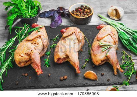 Three singed raw uncooked quail on a cutting board. Spices and herbs for raw quail thyme rosemary basil arugula onions garlic peppers. Light wood background.