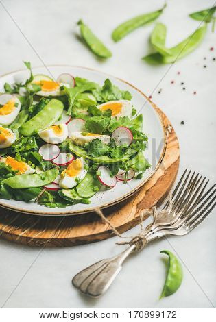 Healthy spring green salad with radish, boiled egg, arugula, green pea and mint in white plate on olive tree wood board over grey background, selective focus, vertical composition