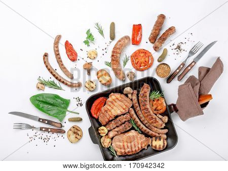Steak and different sausages and grilled vegetables on white background in cast iron grill pan. top view. Flat lay