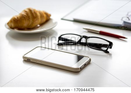 Office breakfast with croissant, office materiales, glasses and mobile phone on white desk