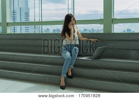 Beautiful Young Asian business women using smartphone and laptop computer sitting on the stairs with happiness and concentrate with city view background.