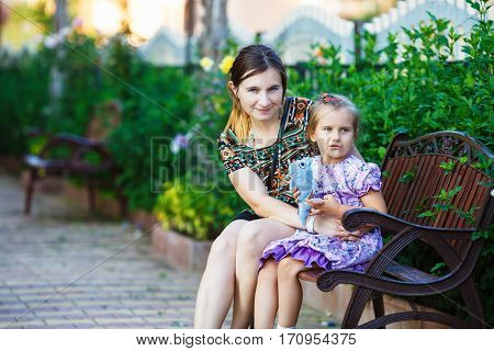 Mother and little daughter sit on a park bench on a background of bright green foliage. Hugging mother and daughter. Family portrait. Selective focus.