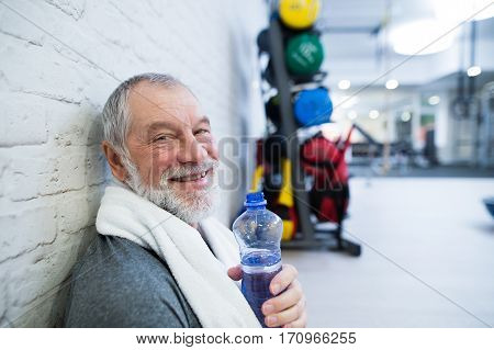 Fit senior man in sports clothing in gym resting after working out, towel around his neck, holding a watter bottle