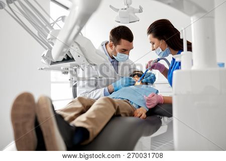 medicine, dentistry and healthcare concept - dentist and assistant with dental drill and saliva ejector treating kid patient teeth at dental clinic