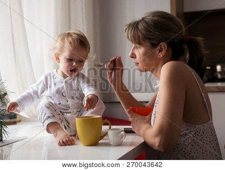 Mom In The Kitchen Feeding The Child With A Spoon. Breakfast. The Child Sits On The Table.