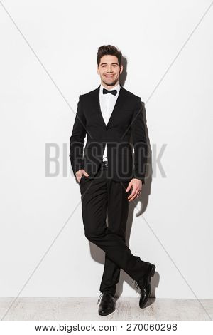 Full length portrait of a smiling young man wearing black suit with a bow standing isolated over white background, posing