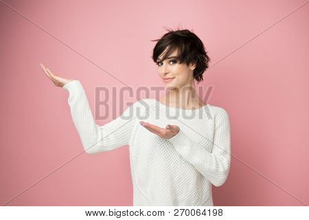 Charming stylish young woman pointing at copy space and smiling at camera. Cute girl displays something over soft pink background.