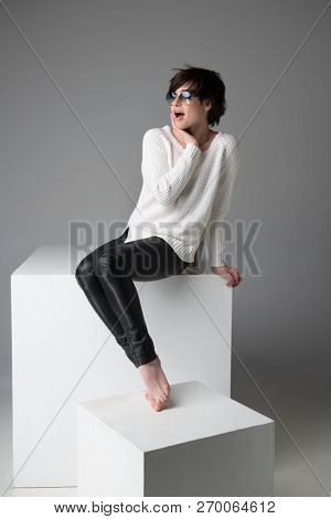 Excited young woman with sunglasses sitting on white cube in studio isolated over gray background, looking at camera. Trendy girl with surprised face expression.
