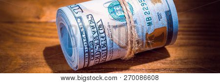 A Roll Of Dollars On A Wooden Textured Table In The Dark Illuminated By A Ray Of Light.
