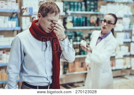Illness Man Have A Head Pain. Pharmacist Is Looking On Man. Man Wearing A Scarf. Pharmacist Is A Bea