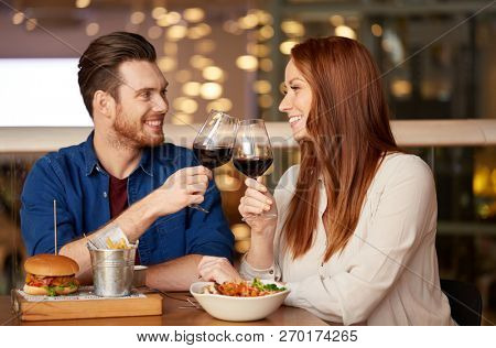 celebration, leisure and holidays concept - happy young couple eating and clinking glasses of red wine at restaurant