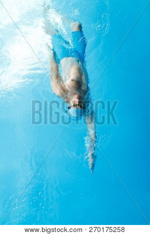 Image on top of young sportsman in blue cap swimming on back in swimming pool