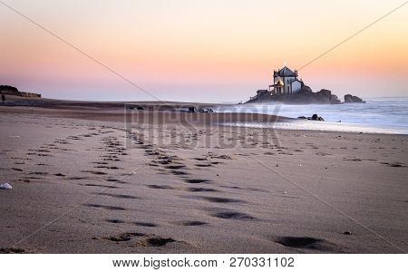 Footsteps Marked On The Sand Lead To The Senhor Da Pedra Chapel At Sunset With Waves Crashing In The