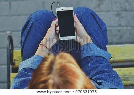 A Young Pretty Student Uses A Smartphone Sitting On A Yellow Bench In A Denim Jacket On The Street