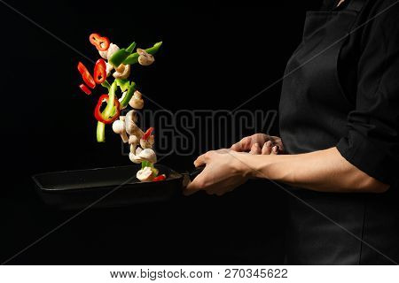 Professional Cook. The Chef Prepares A Dish With Vegetables And Mushrooms In A Saucepan. On A Black
