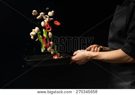 Professional Cook. The Chef Prepares A Dish With Vegetables And Mushrooms In A Saucepan. On A Black