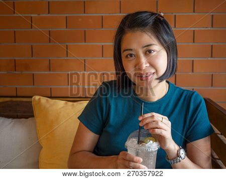 Portrait Of Asian Middle Aged Woman Looking At Camera, Holding Ice Berverage.  Brick Wall, Sitting O