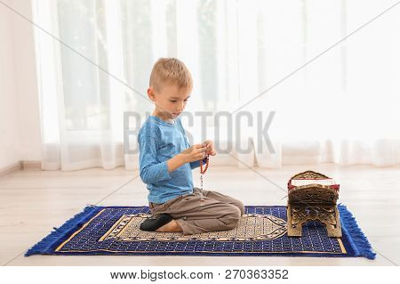 Little Muslim Boy With Misbaha And Koran Praying On Rug Indoors
