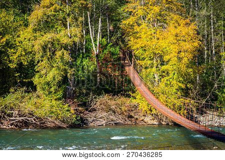 Narrow Metal Foot Bridge Across Mountain River In Autumn.
