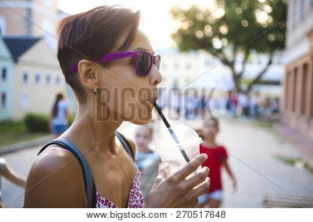 A Girl Drinks A Drink Through A Straw. A Woman In Glasses Drinks Morning Coffee On A City Street. A 