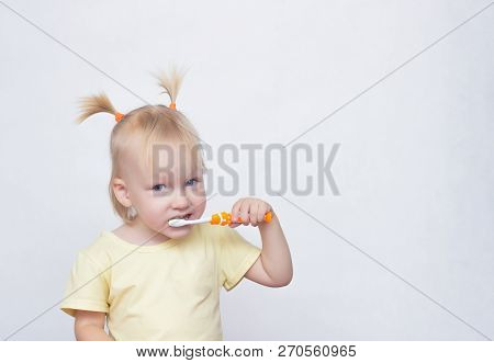 Little Blue-eyed Girl Blonde With Pigtails On Her Head Brushing Teeth With A Toothbrush And Looking 
