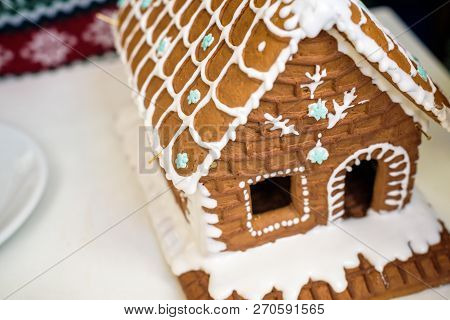 Homemade Christmas Gingerbread House In The Snow. Decorated Christmas Tree In The Background