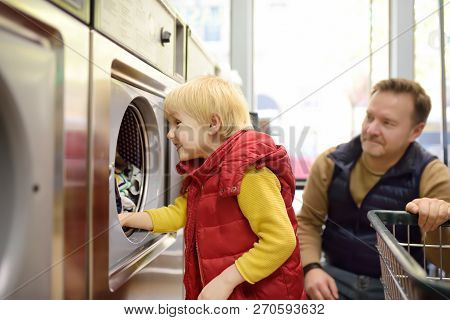A Little Boy Loads Clothes Into The Washing Machine In Public Laundrette. Family In Public Laundry
