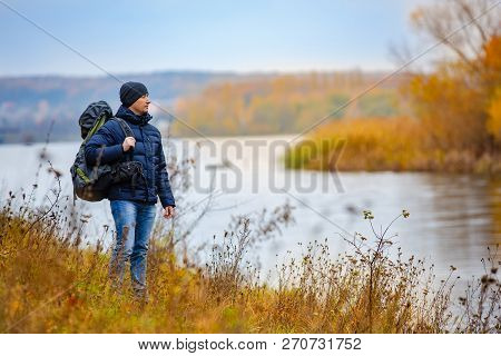 A Healthy Man With A Black Rucksak On The Background Of Blue Sky. Autumn Scenery Near The Calm River