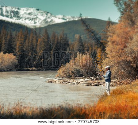 Young amateur angler fishing in the rapid river with snow caped mountains on the background. Altai, Russia. Version of the file with tilt shift effect applied.